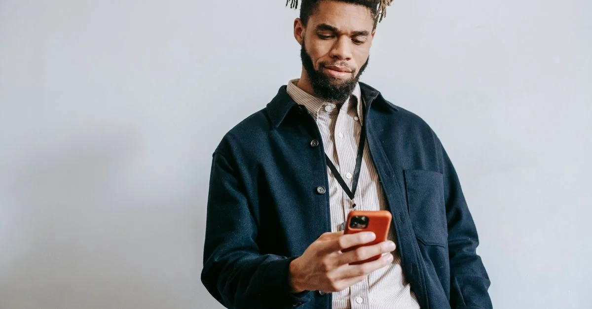 Joven carismático con barba navegando por su smartphone en interiores, mostrando un estilo de vida moderno.
