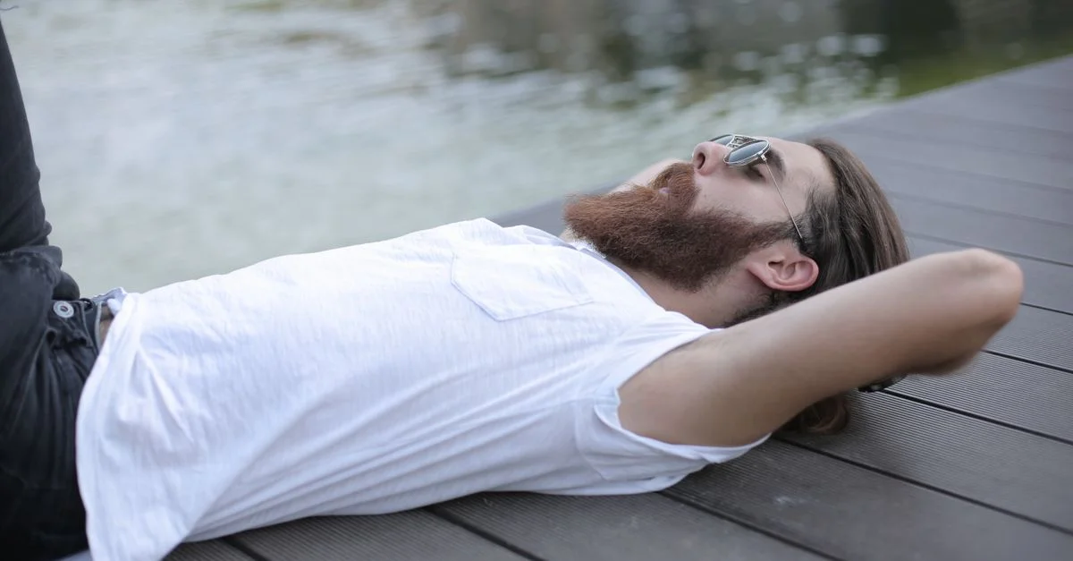 Hombre barbudo en camisa blanca acostado relajado en un muelle junto al lago bajo la luz del día.