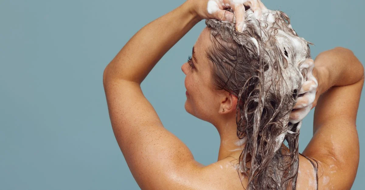 Mujer con cabello largo lavándolo con champú, creando espuma, en un entorno interior contra un fondo azul.